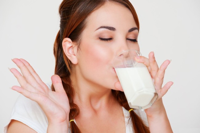 щастлива млада жена, която пие мляко над сивия фон